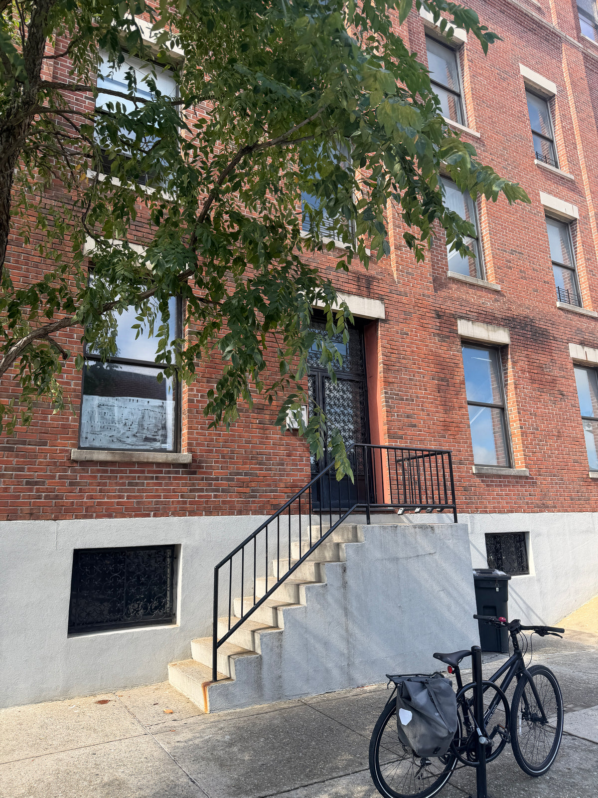 A red brick apartment building with a concrete stairway and black metal railing, photographed outdoors on a sidewalk with a bicycle in the foreground and tree branches overhead.