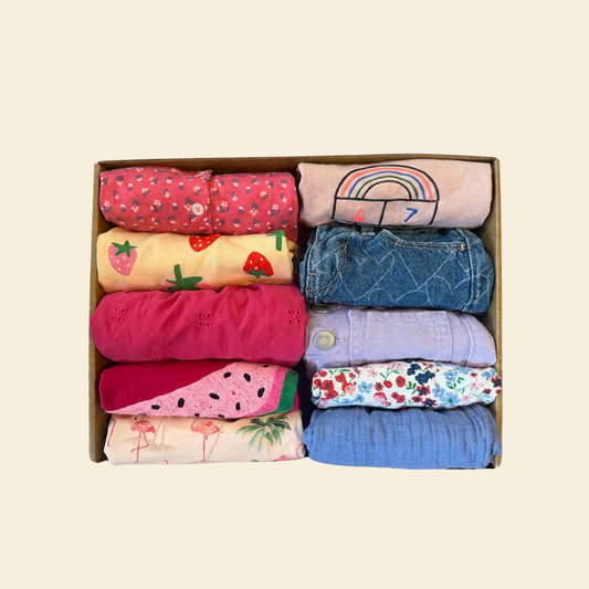 Box of gently used toddler girl clothing featuring pick, blue and purple dresses, shirts and shorts with fruit and flower accents.