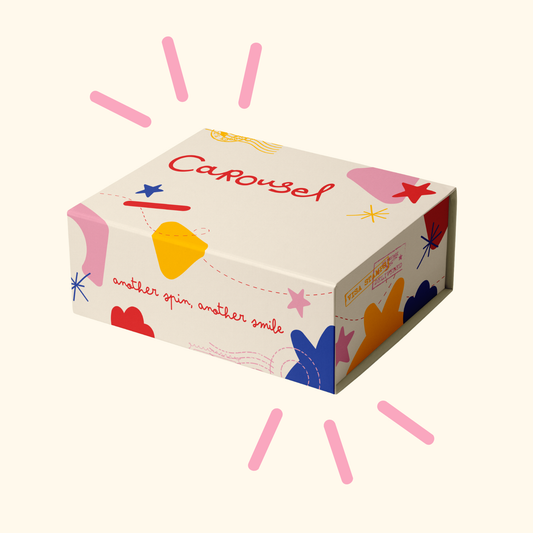 A rectangular product box with a cream-colored background and colorful abstract shapes, featuring the word 'Carousel' in red script on top. The box is shown on a light cream background with pink radiating lines around it.