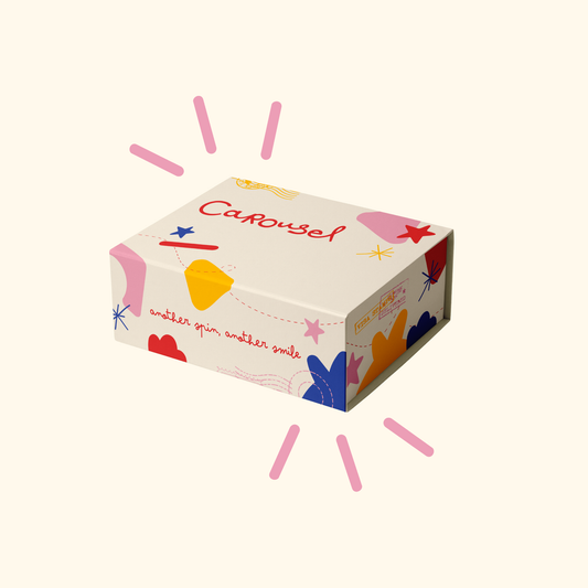 A rectangular product box with a cream background and colorful abstract illustrations, including the word 'Carousel' in red script. The box is shown on a light cream background with decorative pink radiating lines around it.