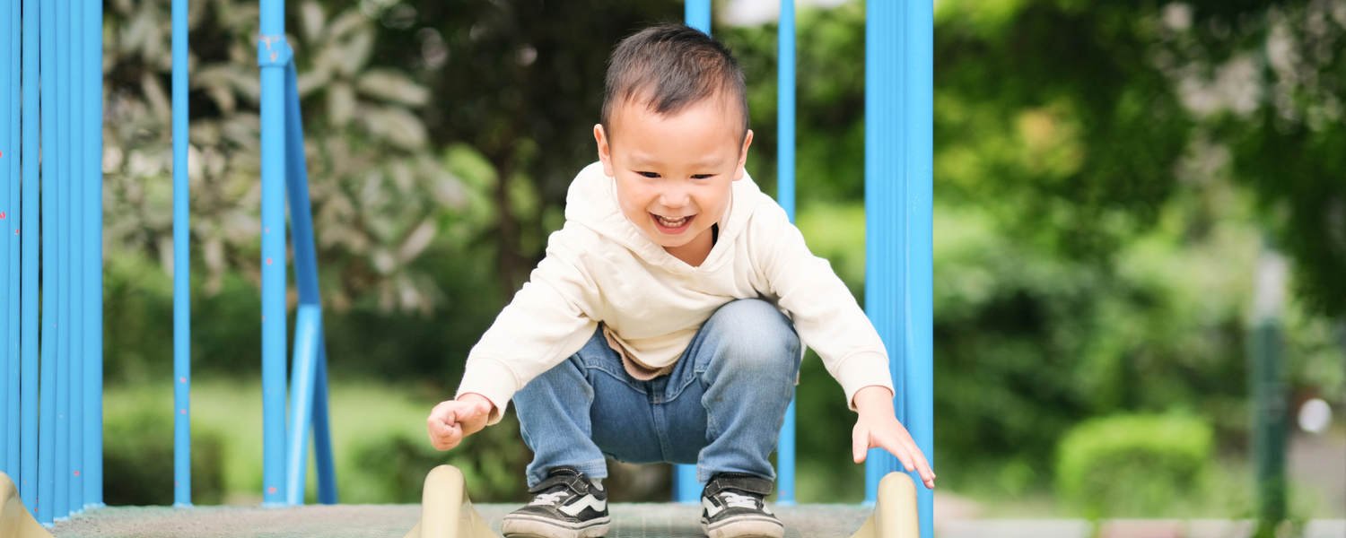 A young toddler boy smiling and crouching on a playground slide, wearing a cream long-sleeve shirt, blue jeans, and black sneakers, with a blurred green park background and blue playground bars.