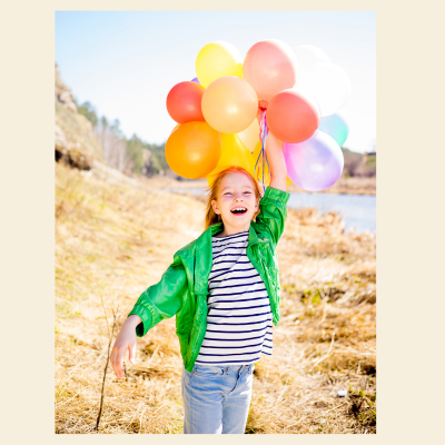 A young girl laughing outdoors while holding a bunch of colorful balloons, wearing a shiny green jacket over a navy and white striped shirt and light blue jeans.