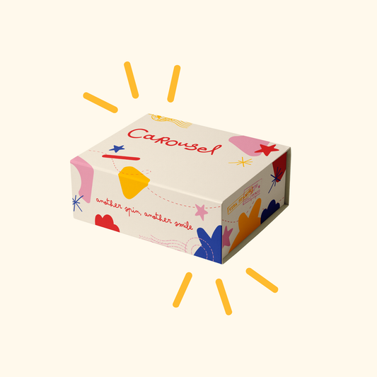 A rectangular product box with a cream background and colorful abstract shapes, including the word 'Carousel' in red script. The box is shown floating on a light cream background with yellow sunburst lines around it.