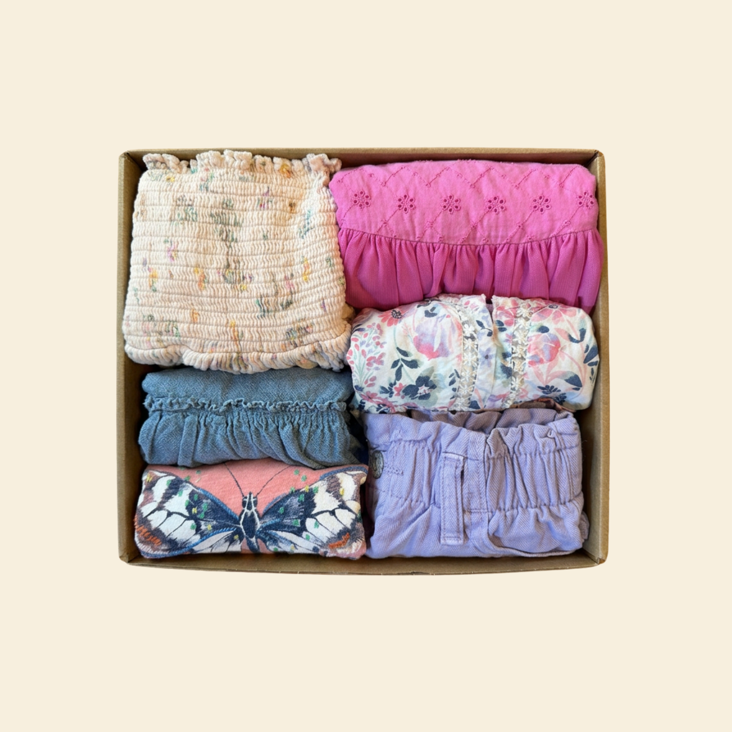 A cardboard gift box containing six folded baby clothing items in various prints and colors, including floral, butterfly, and solid ruffled fabrics, set against a solid light beige background.