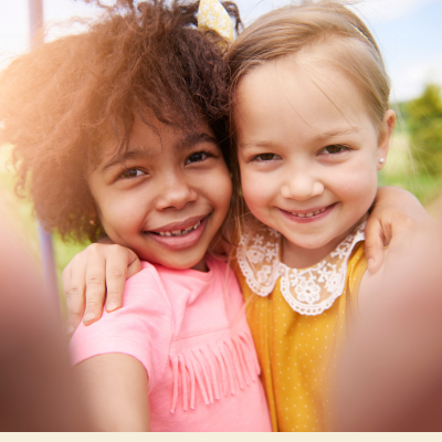 Two young girls smiling outdoors, one wearing a pink dress with fringe and the other wearing a yellow top with a white lace collar, with a soft-focus blurred background.