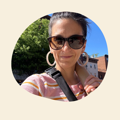 Woman wearing sunglasses and large hoop earrings outdoors with trees and buildings in the background.